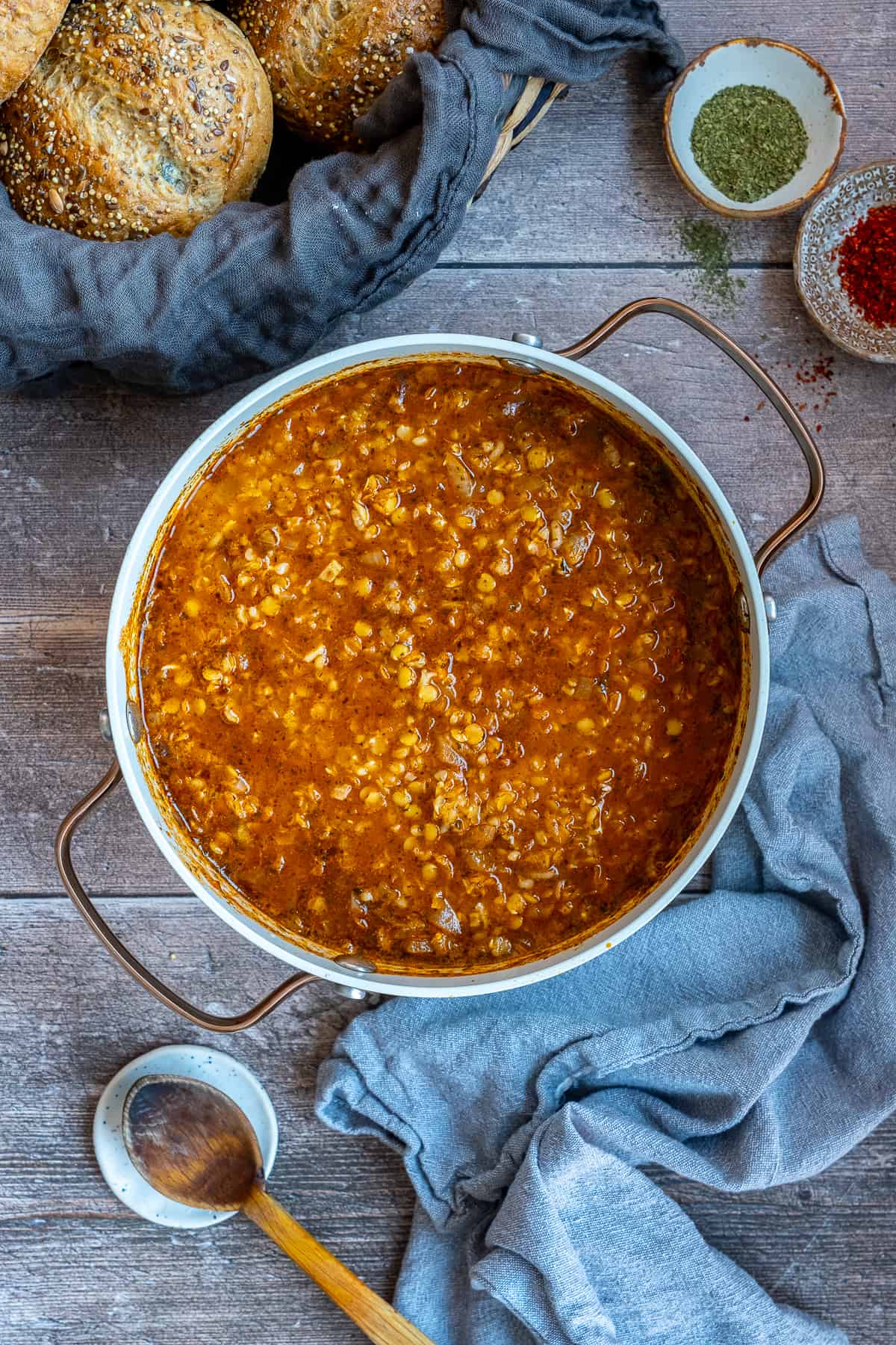 Ezogelin soup in a white pot from top view, a wooden spoon, spices and bread rolls accompany.