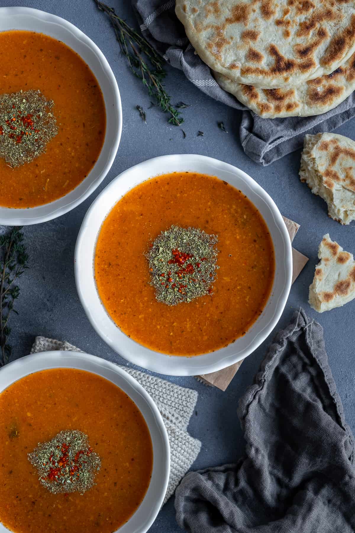 Tarhana soup in three white bowls, garnished with dried mint and red pepper flakes and some Turkish flatbread accompanies.
