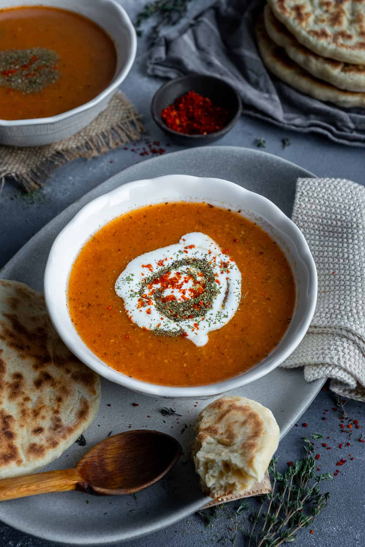 Tarhana soup topped with yogurt, dried mint and red pepper flakes in a white bowl, some flat bread and a wooden spoon accompany.