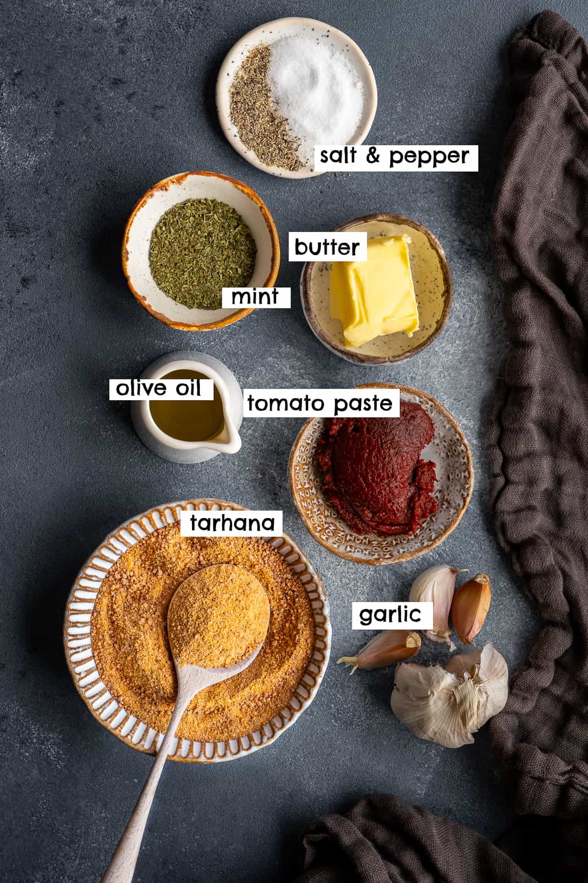 Tarhana soup powder, tomato paste, butter, olive oil, garlic cloves, dried mint, salt and pepper on a grey background.