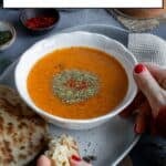 A bowl of tarhana soup garnished with dried mint and red pepper flakes, some bread and a wooden spoon on the side.