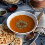 A bowl of tarhana soup garnished with dried mint and red pepper flakes, some bread and a wooden spoon on the side.