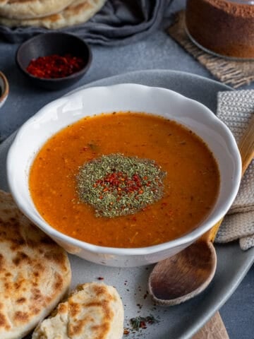 A bowl of tarhana soup garnished with dried mint and red pepper flakes, some bread and a wooden spoon on the side.