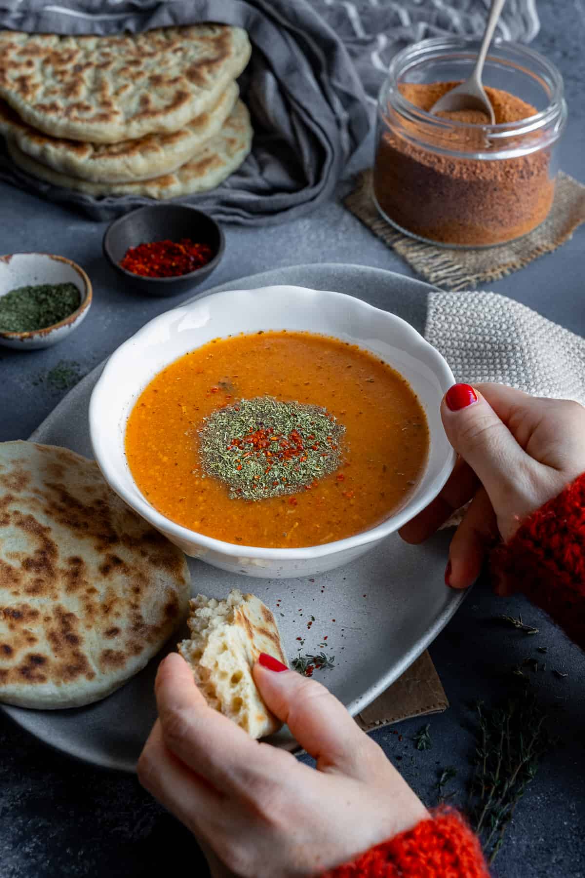 Woman hands holding a bowl of soup and some piece of flatbread accompanied by spices and Turkish tarhana in a jar.