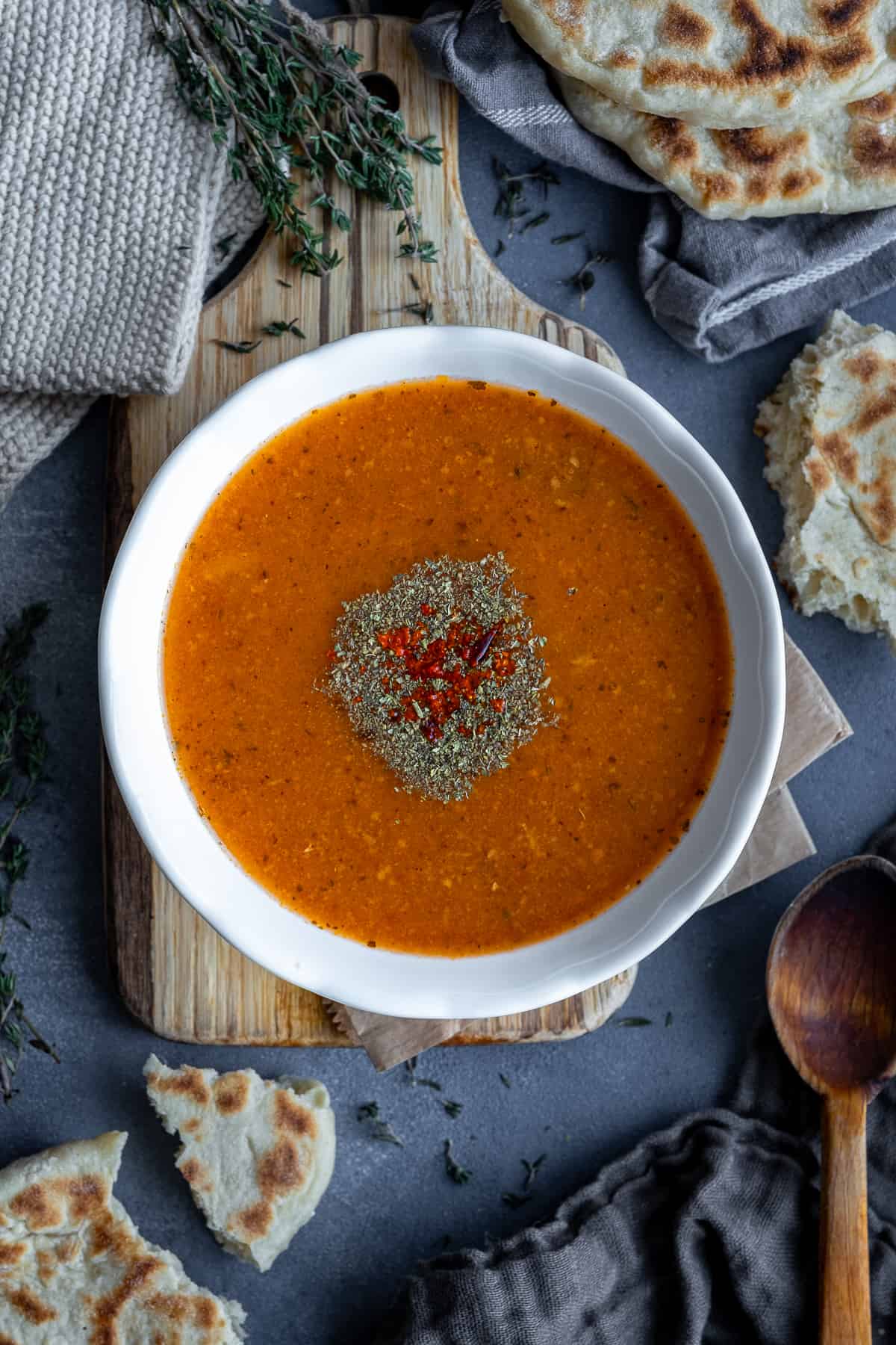 Turkish tarhana soup in a white bowl, garnished with dried mint and red pepper flakes, Turkish bazlama bread, thyme and a wooden spoon accompany.