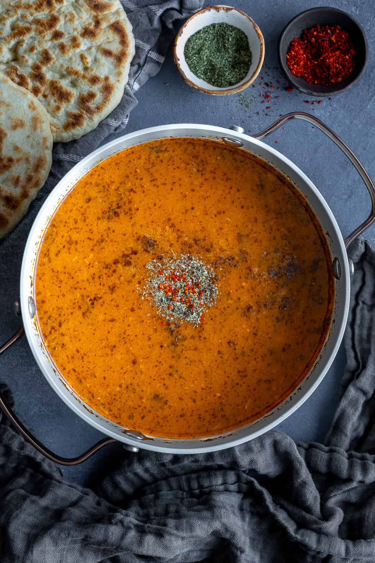 Tarhana soup in a white pot accompanied by Turkish bazlama bread, dried mint and red pepper flakes.
