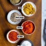 Oregano, cumin, black pepper, paprika, red pepper flakes and salt each in mini bowls on a wooden board.