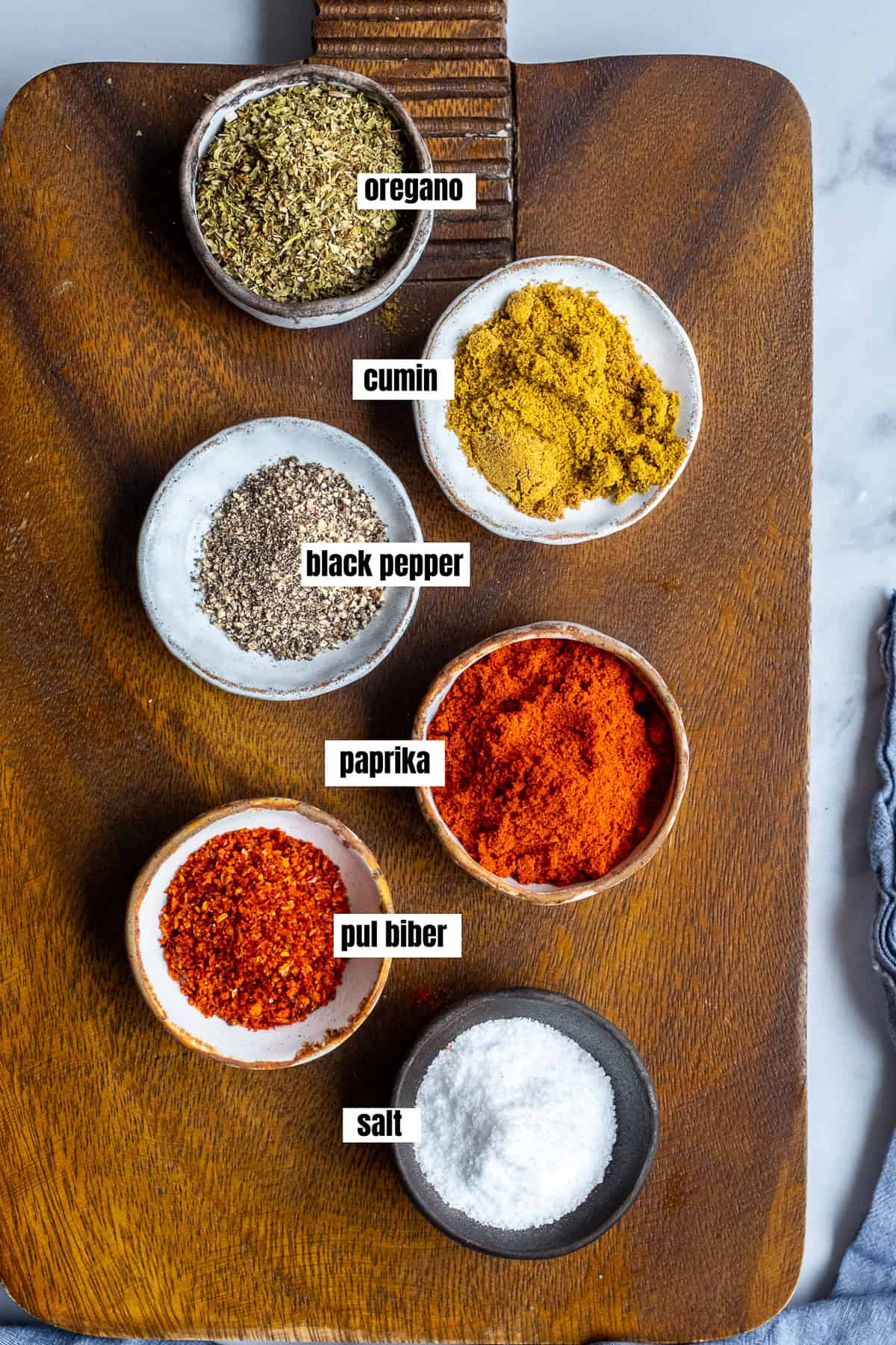 Oregano, cumin, black pepper, paprika, red pepper flakes and salt each in mini bowls on a wooden board.