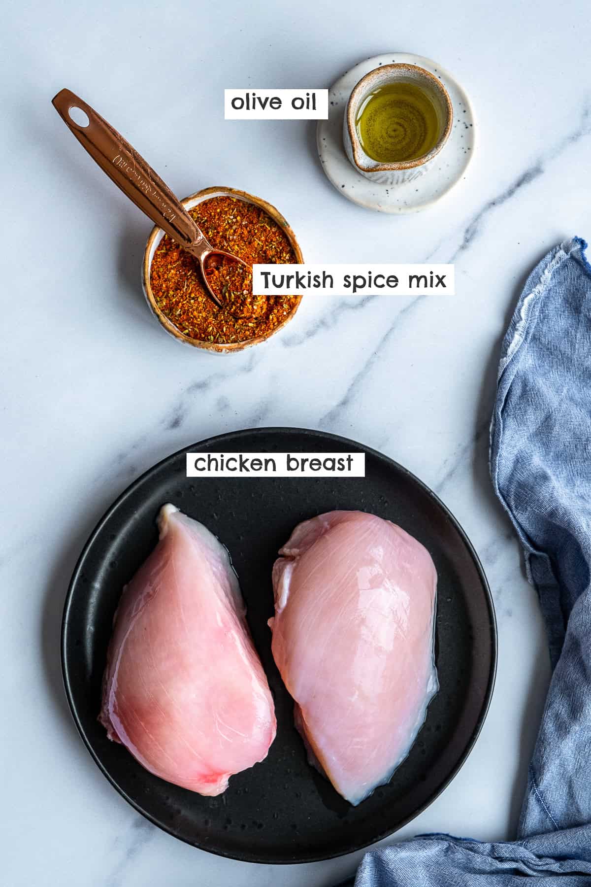 Uncooked chicken breasts on a black plate, spice mix in a small bowl and olive oil in a mini jug on a white marble.