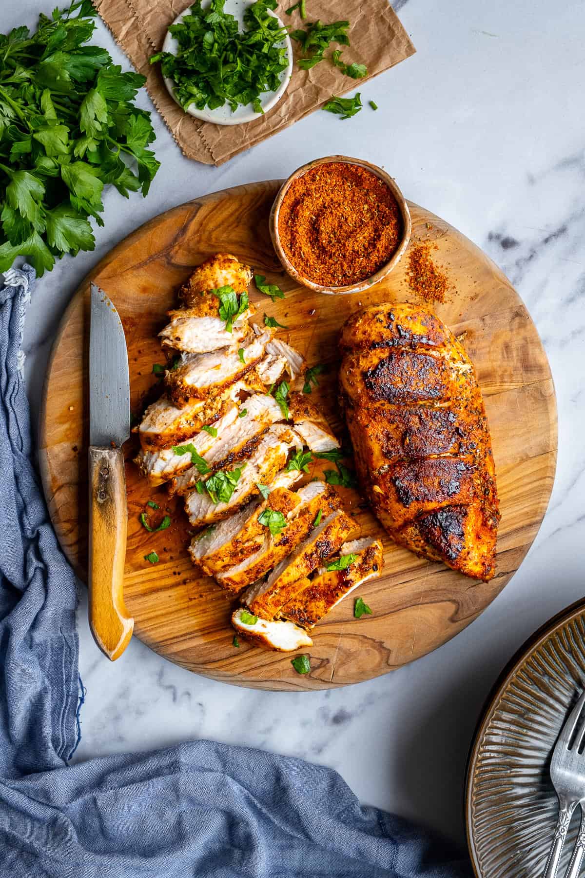 Pan-fried chicken breasts  on a round wooden cutting board, one is sliced and a spice mix in a small bowl on the side.