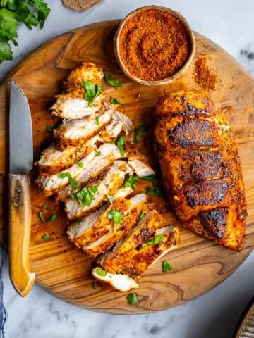 Pan-fried chicken breasts on a round wooden cutting board and a spice mix in a small bowl on the side.