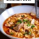 Chicken soup with a tomato base and orzo served in a white bowl and a spoon inside it.