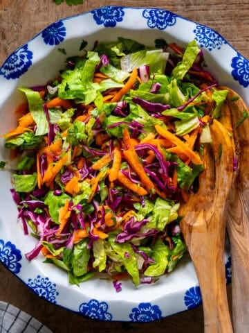 A fresh salad with lettuce, carrots and red cabbage in a white bowl and wooden spoons inside it.