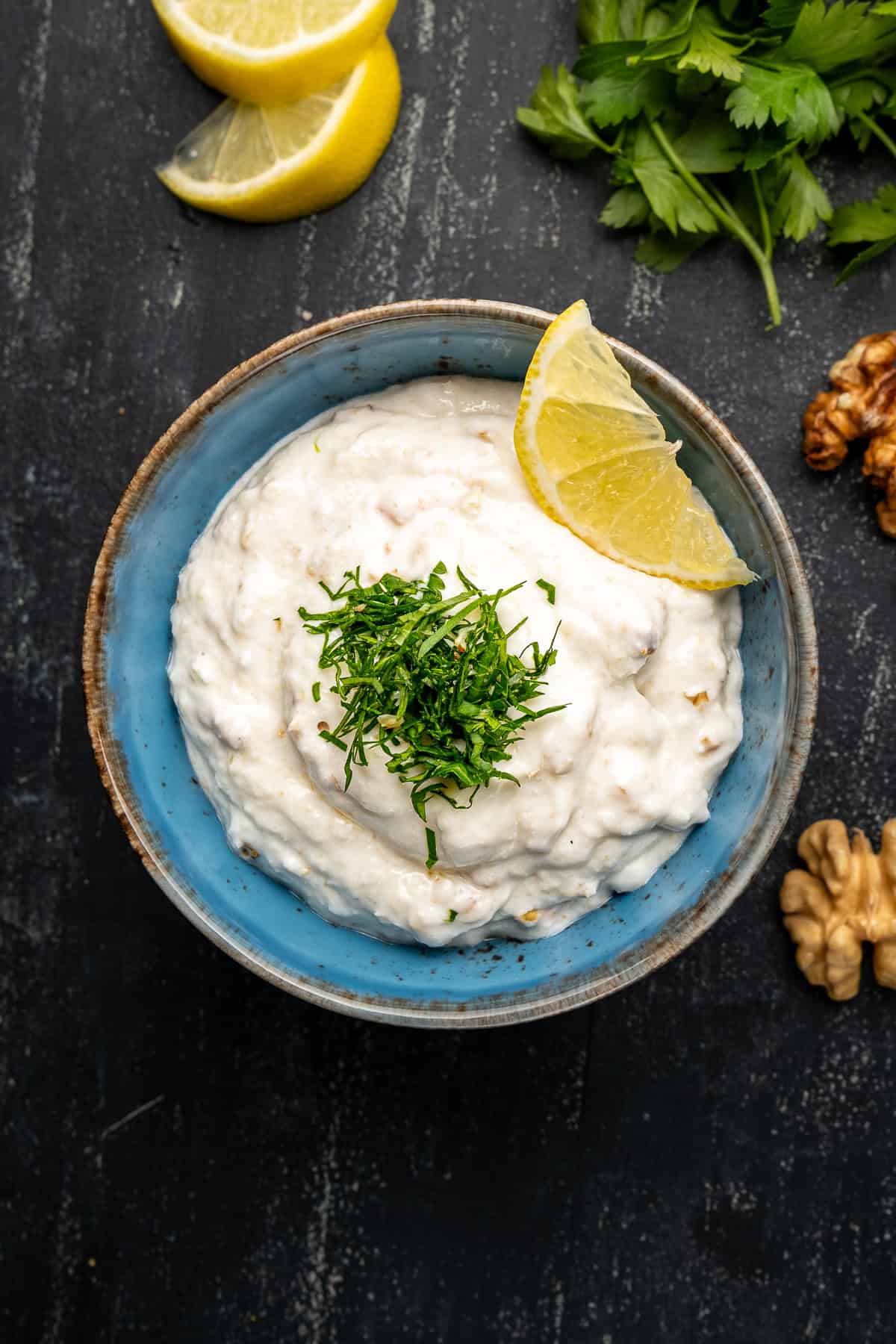 Tarator sauce with yogurt, breadcrumbs and walnuts garnished with chopped parsley and a slice of lemon in a blue bowl.