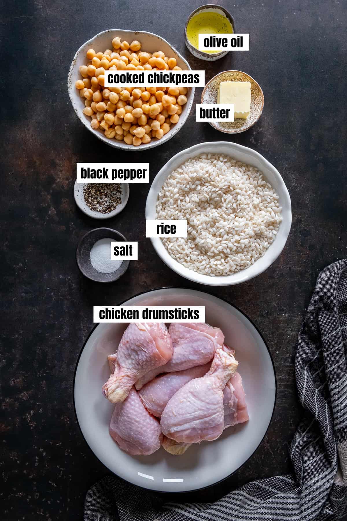 Labeled ingredients for Tavuklu Pilav, including chicken drumsticks, rice, cooked chickpeas, butter, olive oil, salt, and black pepper, arranged on a dark surface.