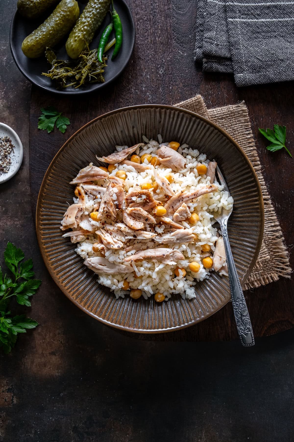 A bowl of Tavuklu Pilav (Turkish rice with chicken and chickpeas), served with a fork, pickles, and herbs on a rustic wooden surface.