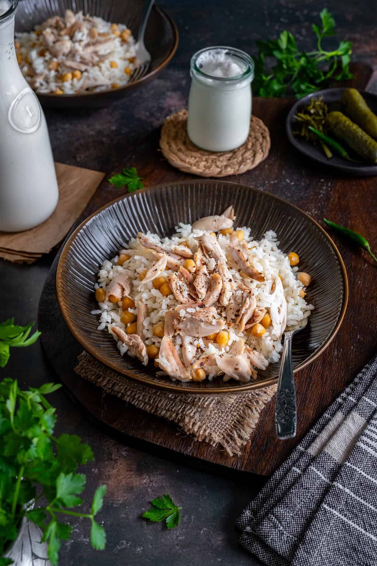 A bowl of Tavuklu Pilav (Turkish rice with chicken and chickpeas) served with ayran, pickles, and herbs on a rustic table setting.