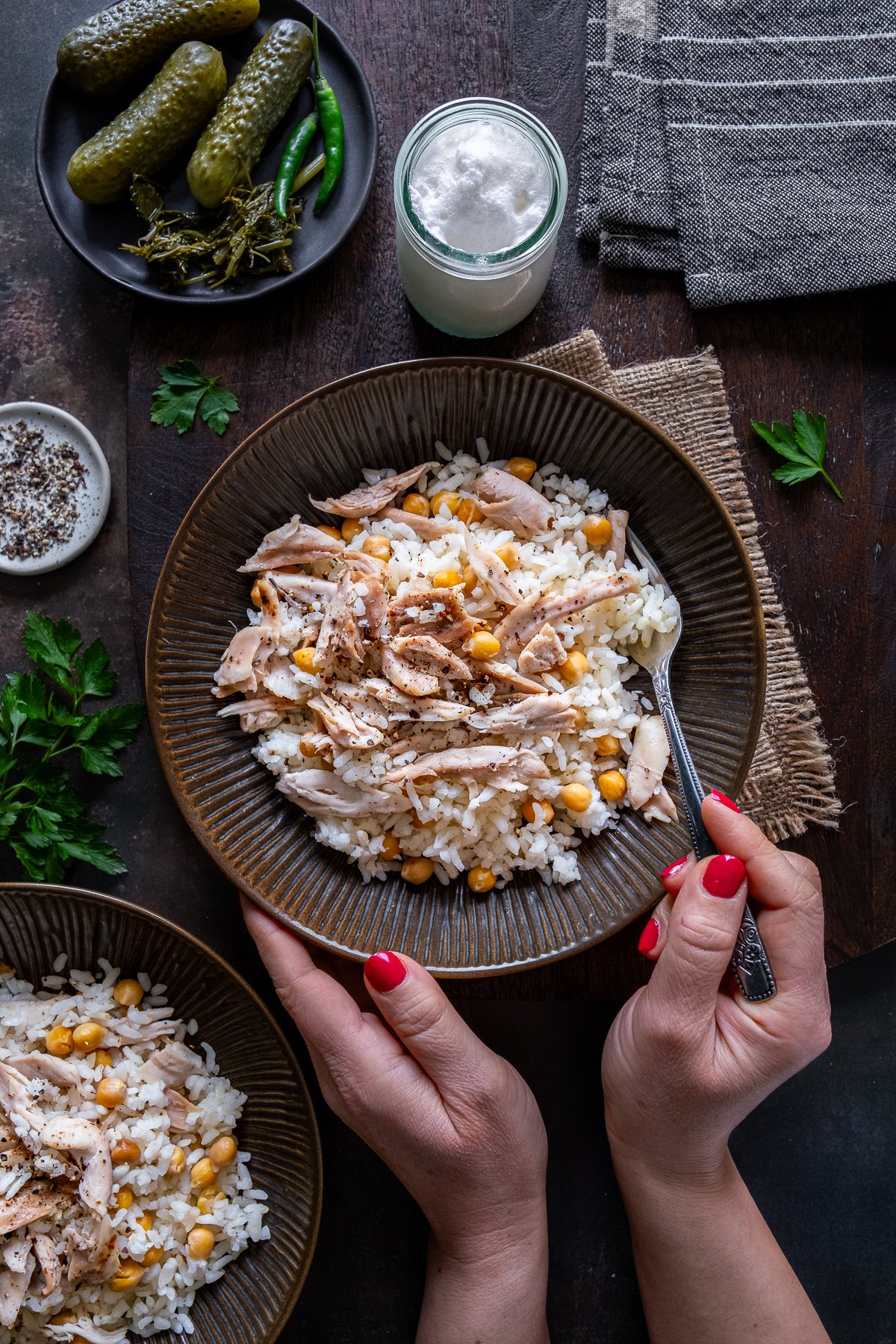 Hands holding a bowl of Tavuklu Pilav (Turkish rice with chicken and chickpeas), served with black pepper, pickles, and a glass of ayran on a rustic table.