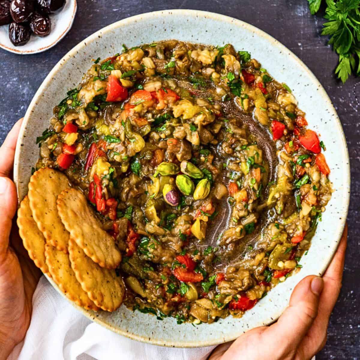 A bowl of chunky baba ganoush without tahini, made with roasted eggplant, tomatoes, herbs, and topped with pistachios, served with crackers.