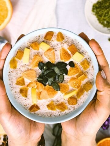 Bulgur breakfast bowl topped with orange, dried fruit, diced quince, and pumpkin seeds, held in hands.