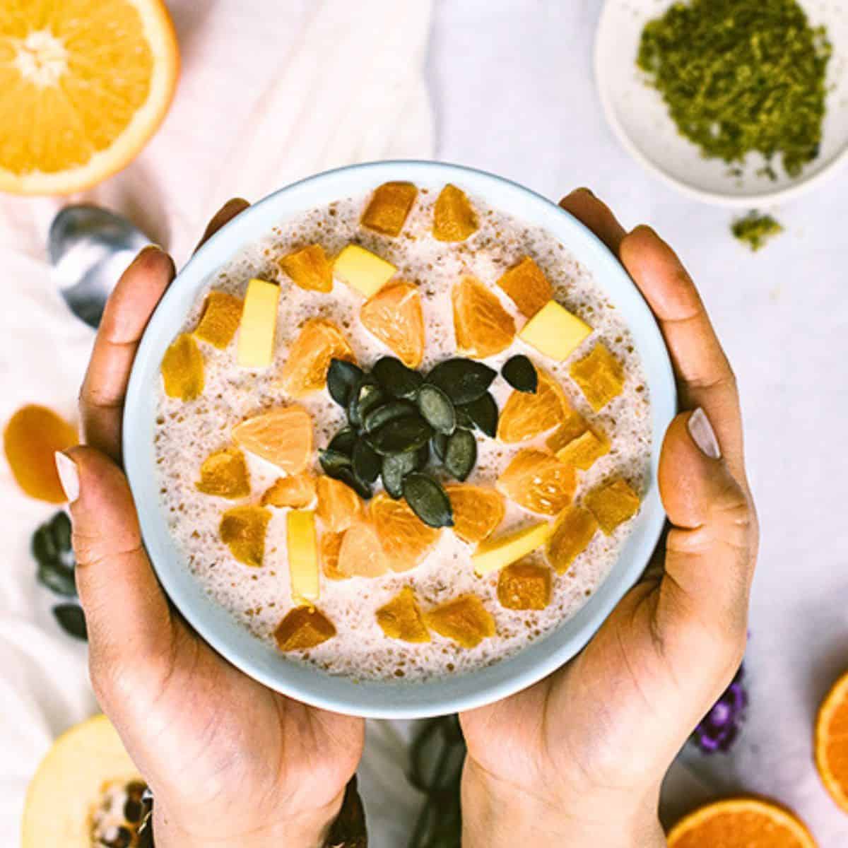 Bulgur breakfast bowl topped with orange, dried fruit, diced quince, and pumpkin seeds, held in hands.