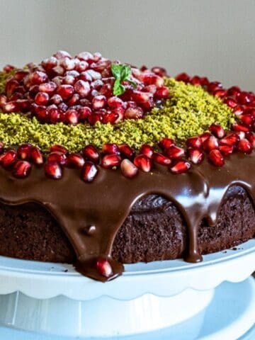 A rich chocolate yogurt cake topped with glossy chocolate ganache, pomegranate seeds, ground pistachios, and a mint garnish, displayed on a white cake stand.