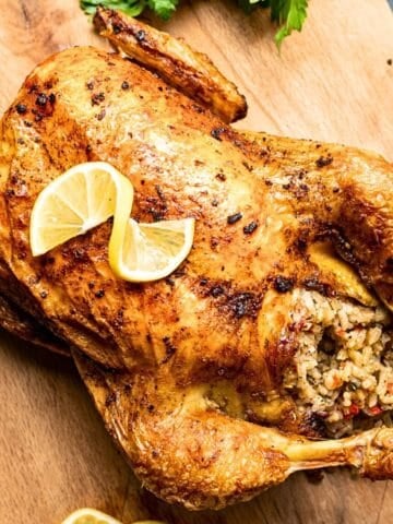 A whole roasted chicken stuffed with savory rice and vegetables, garnished with lemon slices, served on a wooden board.