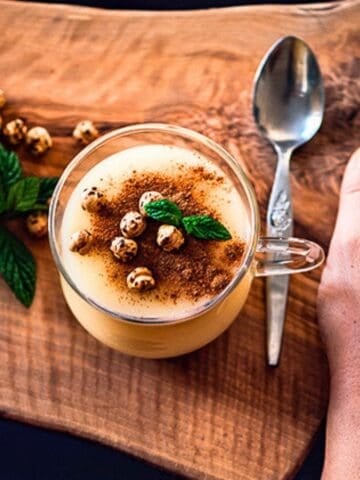 A glass cup of Turkish boza topped with cinnamon, roasted chickpeas, and mint leaves, served on a wooden board with a spoon.