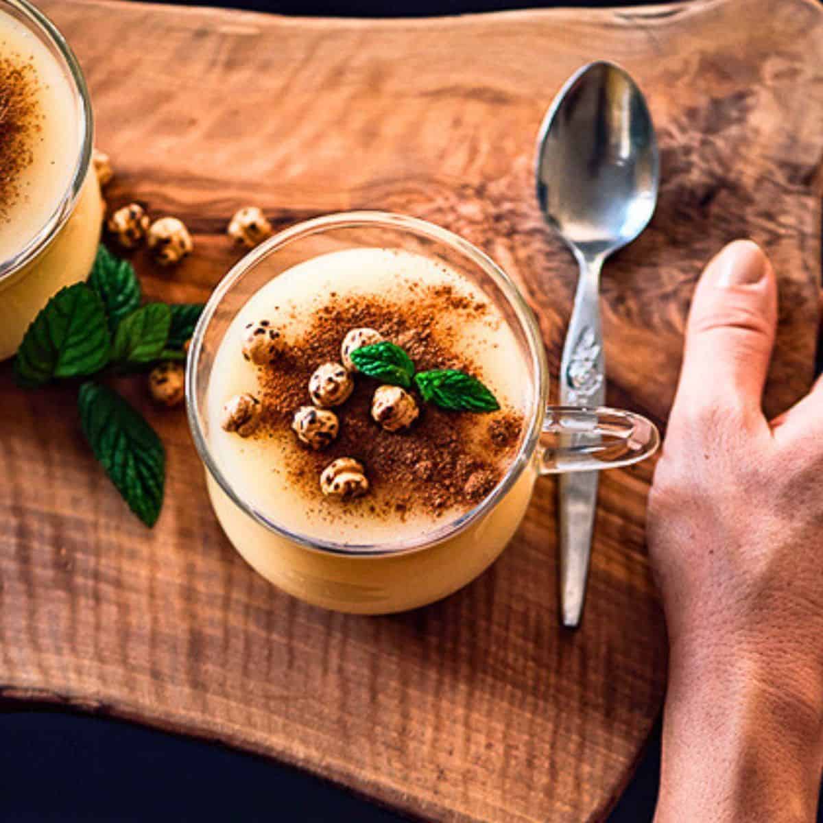 A glass cup of Turkish boza topped with cinnamon, roasted chickpeas, and mint leaves, served on a wooden board with a spoon.