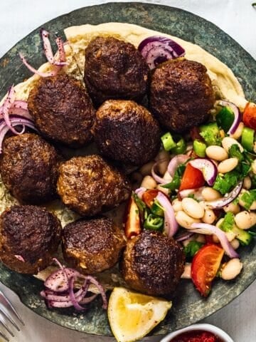 A plate of Turkish meatballs served on flatbread with sliced red onions, a fresh white bean salad with tomatoes and herbs, and a lemon wedge on the side.