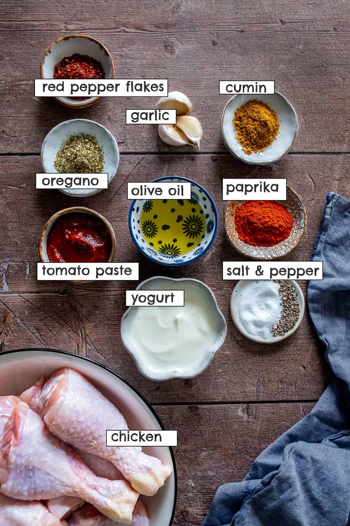 Ingredients for Turkish chicken marinade arranged on a wooden surface, including yogurt, tomato paste, olive oil, garlic, paprika, cumin, oregano, red pepper flakes, salt and pepper, with raw chicken drumsticks in a bowl.