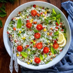 Turkish rice salad in a white bowl, topped with cherry tomatoes, fresh herbs, and lemon slices, with two serving spoons on the side.