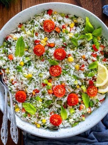 Turkish rice salad in a white bowl, topped with cherry tomatoes, fresh herbs, and lemon slices, with two serving spoons on the side.