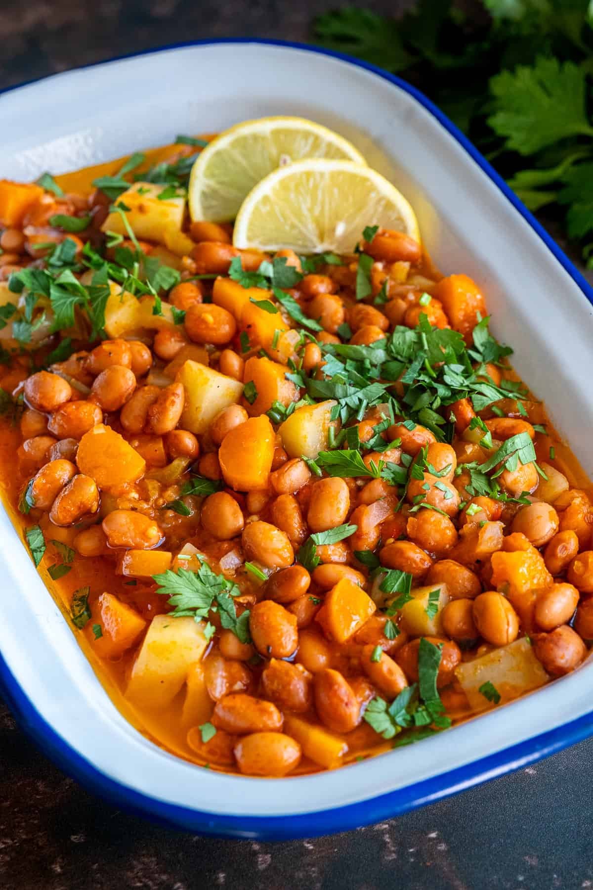 Close shot of Turkish barbunya pilaki in a white enamel dish with carrots, potatoes, and beans, topped with parsley and lemon slices.