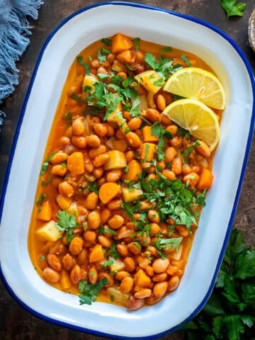 Overhead shot of Turkish barbunya pilaki in a white enamel dish, garnished with parsley and lemon slices.