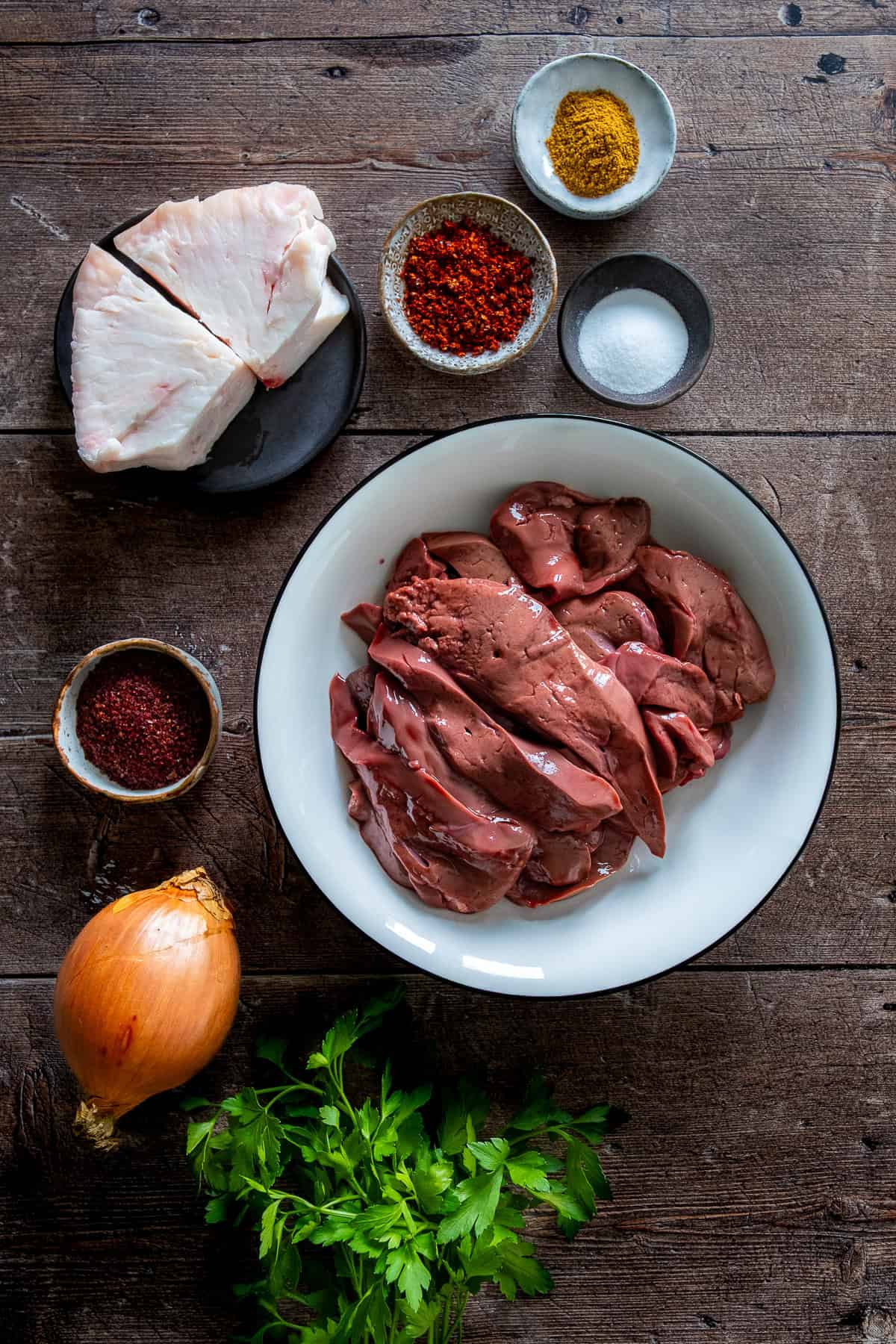 Raw lamb liver, lamb tail fat, onion, parsley, and spices including cumin, salt, pul biber, and sumac arranged on a wooden surface.