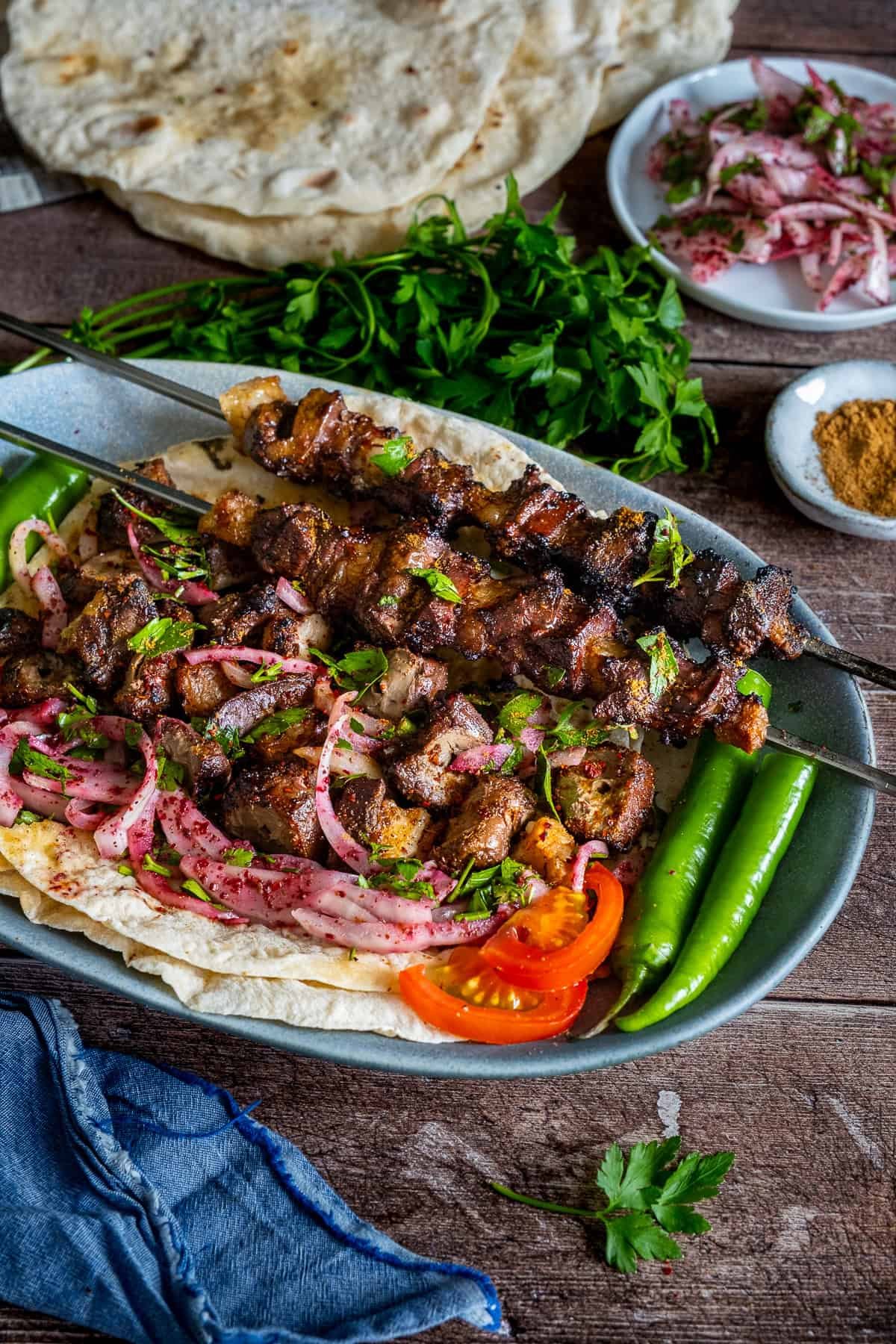 Grilled Turkish liver kebabs on skewers served over lavash bread with sumac onion salad, sliced tomatoes, green peppers, and fresh parsley from front view.