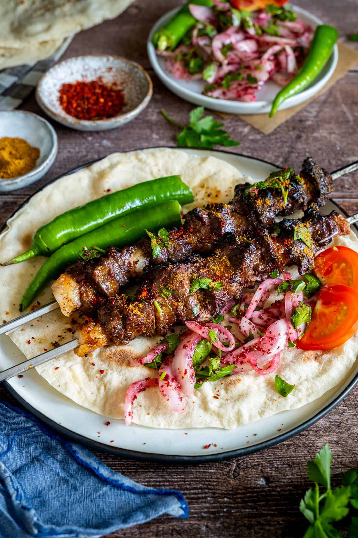 Liver kebabs on skewers served over lavash bread with green peppers, sumac onion salad, sliced tomatoes, and sprinkled spices.