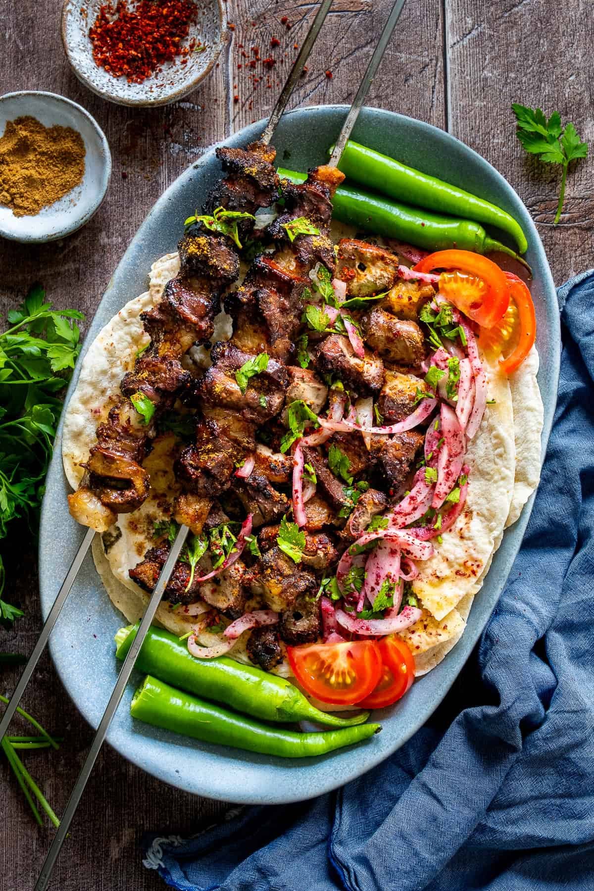 Grilled liver kebabs on skewers served over lavash bread with sumac onion salad, sliced tomatoes, green peppers, and fresh parsley.