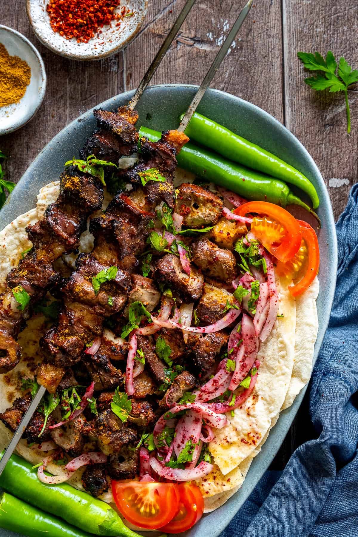 Two skewers of grilled Turkish liver kebabs served on lavash bread with sumac onion salad, fresh parsley, sliced tomatoes, and green peppers, garnished with cumin and pul biber.