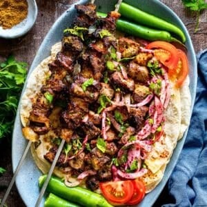 Two skewers of grilled Turkish liver kebabs served on lavash bread with sumac onion salad, fresh parsley, sliced tomatoes, and green peppers, garnished with cumin and pul biber.