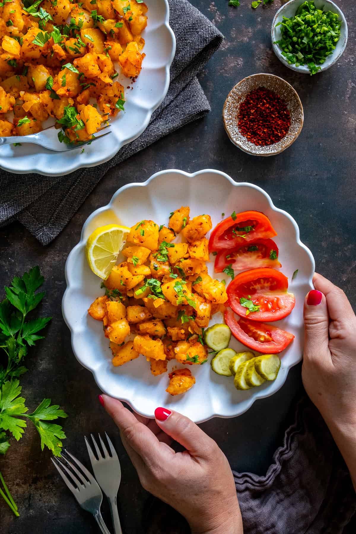 Hands holding a plate of Patates Kavurma with lemon, sliced tomatoes, pickles, and parsley.
