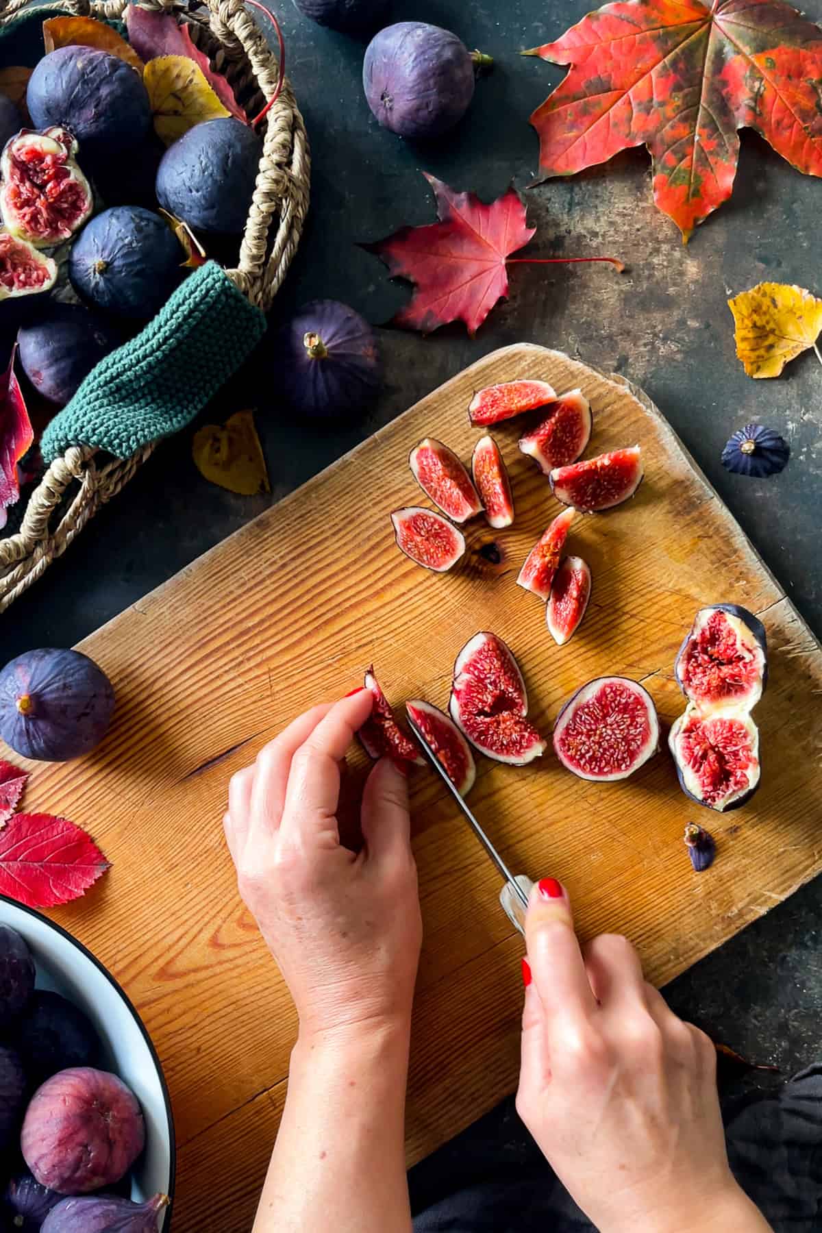 Hands slicing fresh figs on a wooden board with whole figs and autumn leaves around.