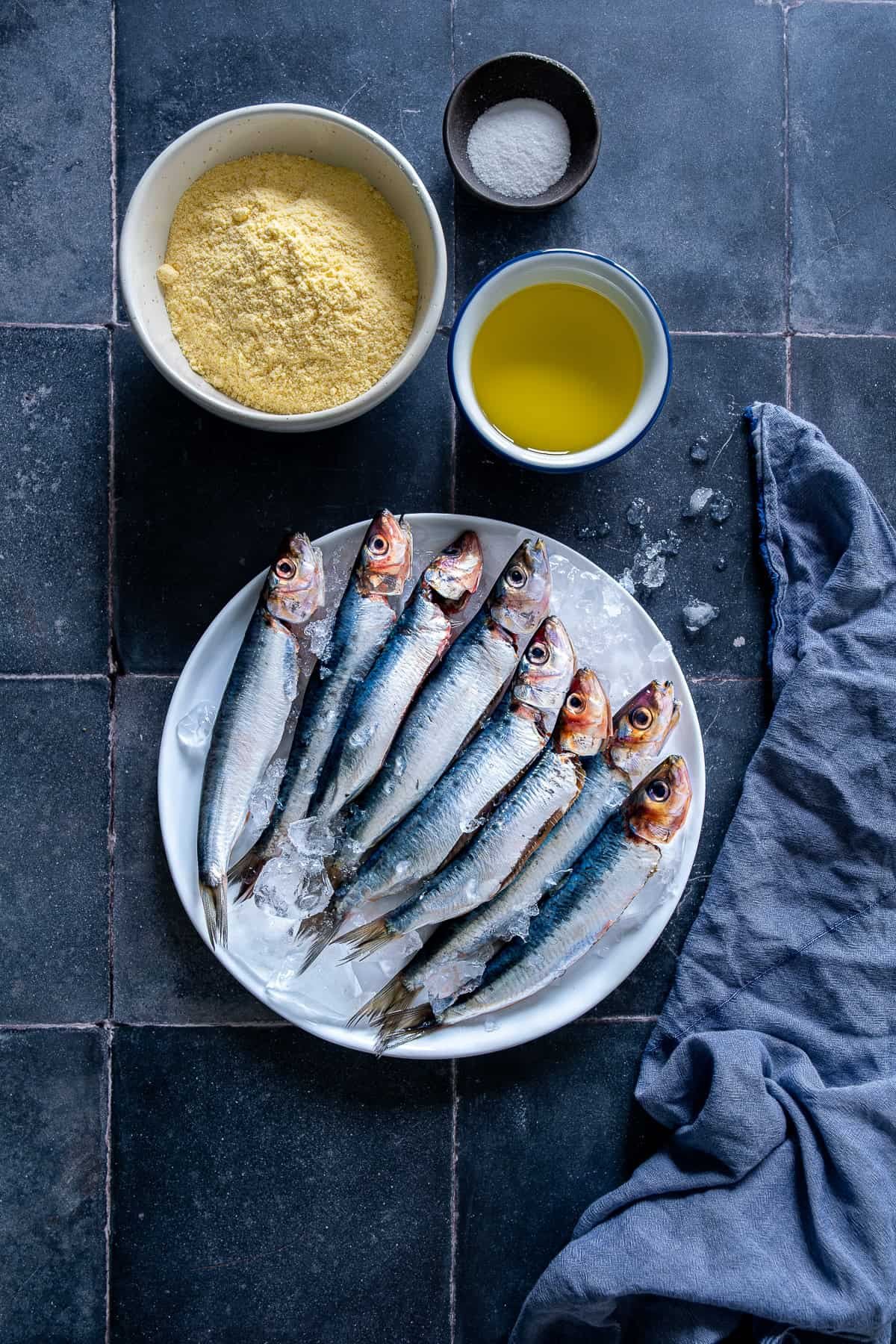 Fresh whole sardines on ice on a white plate, surrounded by cornmeal, salt, and oil on a dark tiled surface.