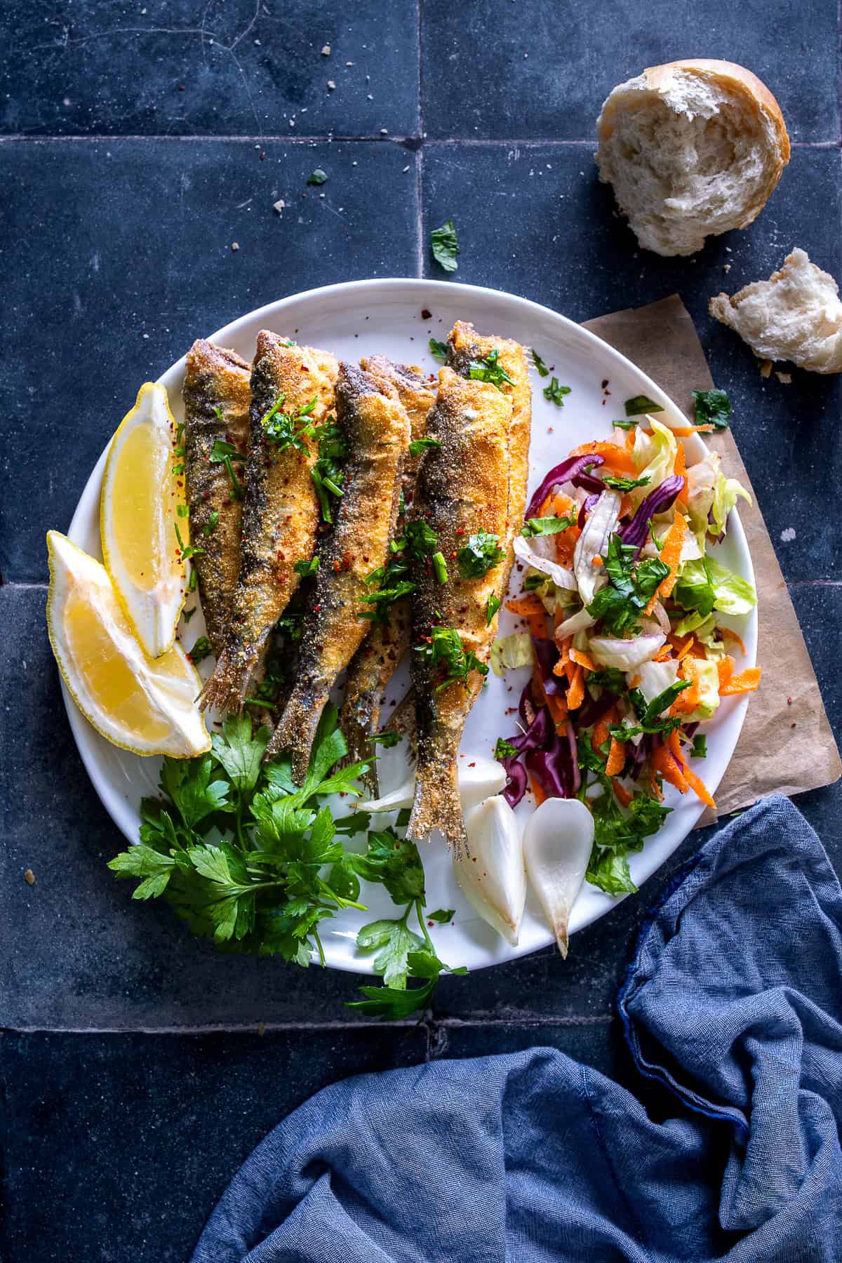 Golden fried sardines served with lemon wedges, fresh parsley, and a colorful salad on a white plate.