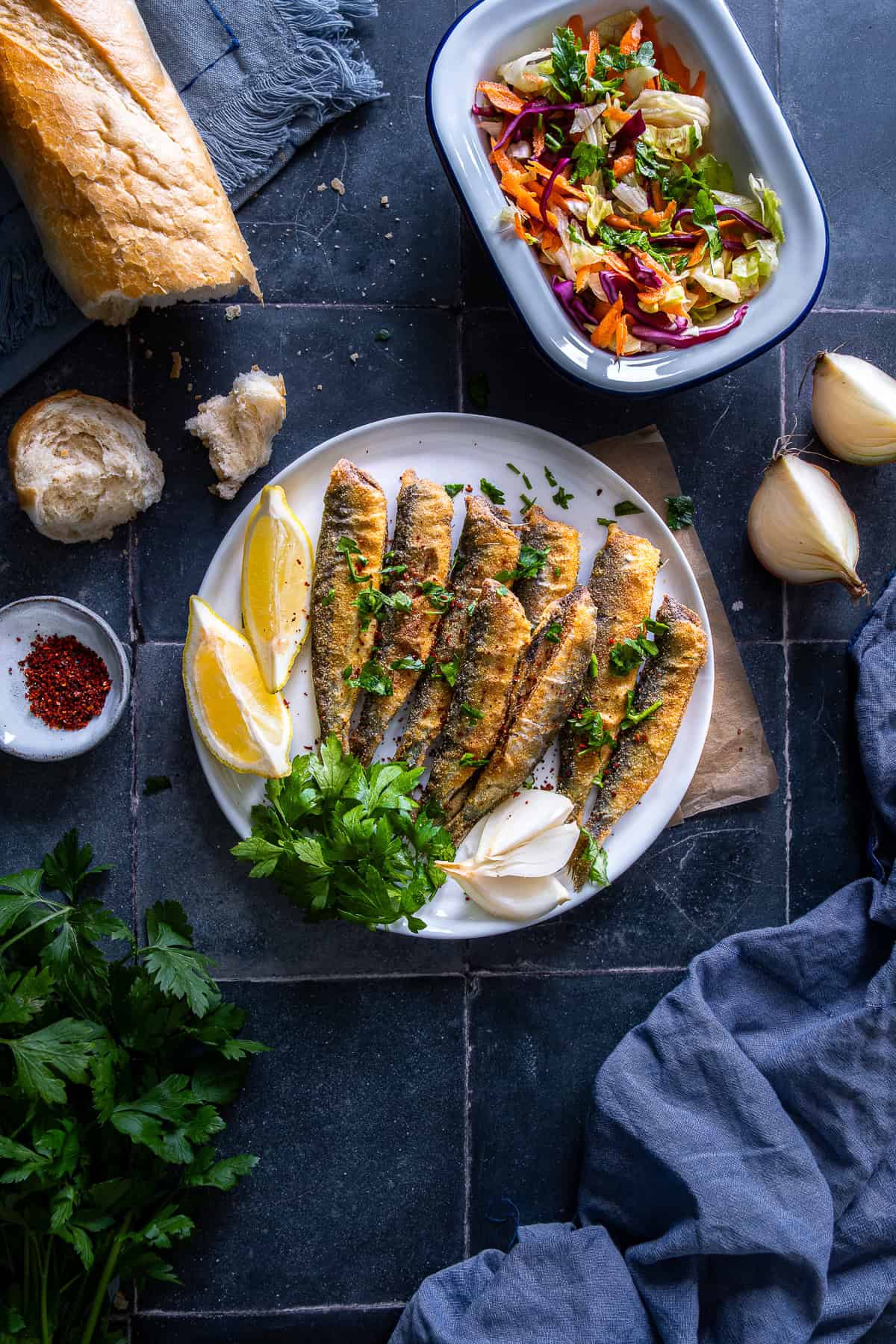 Plate of golden fried sardines with lemon, parsley, and onion, served alongside a fresh salad and crusty bread.