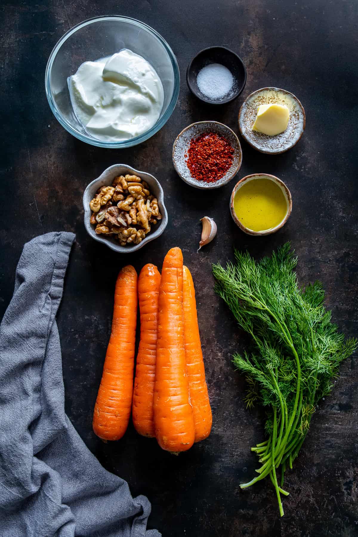 Overhead view of ingredients for Havuç Tarator, including carrots, yogurt, walnuts, dill, garlic, butter, olive oil, pul biber, and salt arranged on a dark surface.