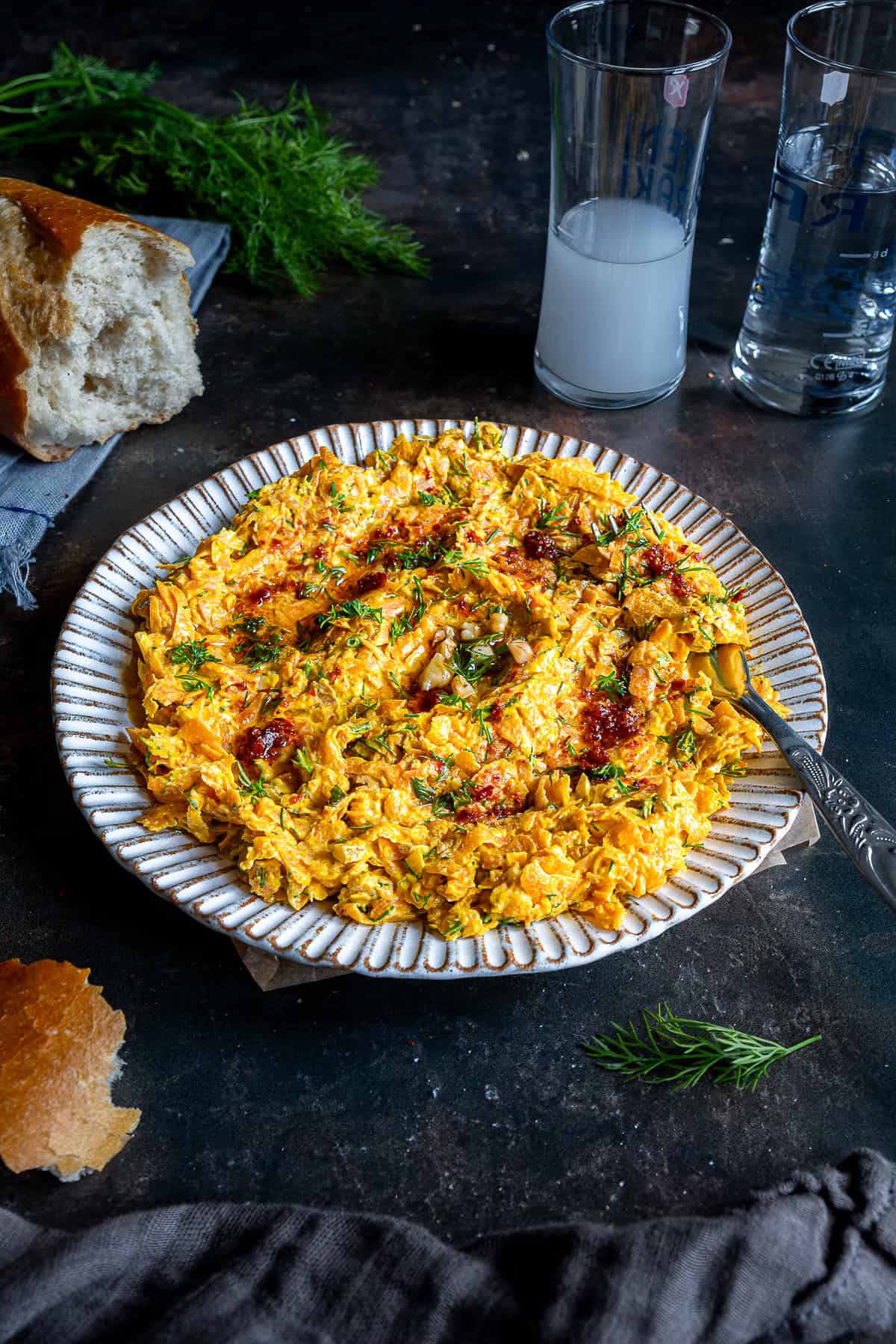 Plate of Havuç Tarator topped with dill and chili oil, served with bread and glasses of rakı on a dark tabletop.
