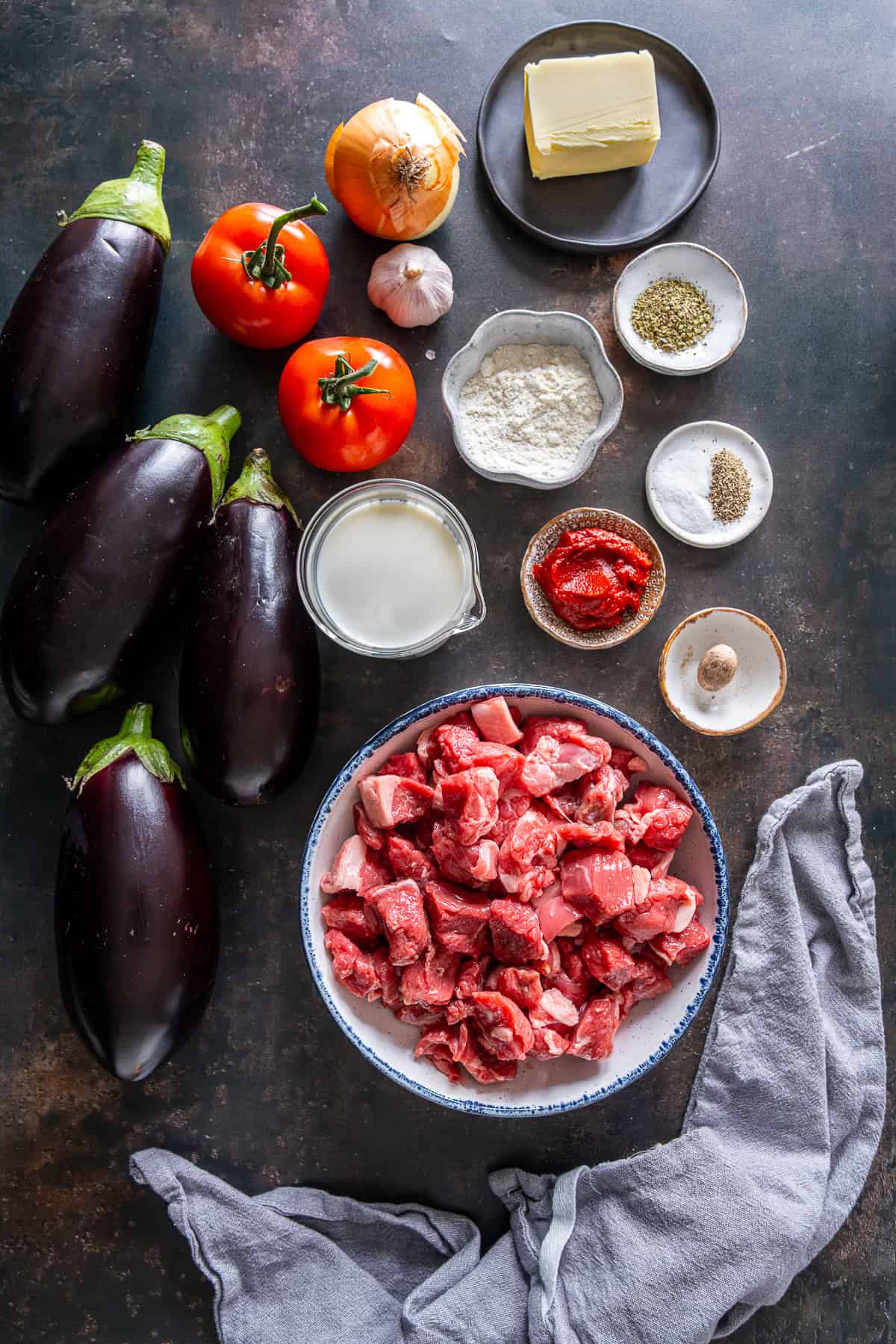 Ingredients for Hunkar Begendi laid out on a dark surface, including eggplants, diced lamb, tomatoes, onion, garlic, milk, butter, flour, and spices.