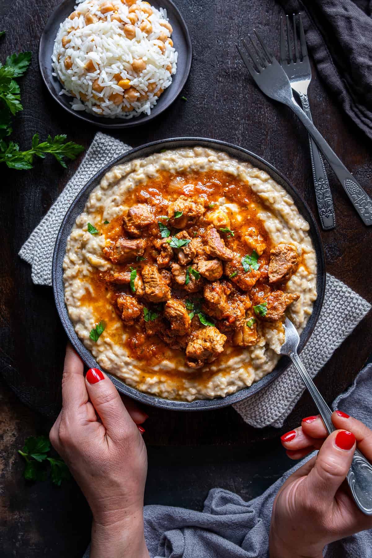 Hands holding a plate of Hunkar Begendi with lamb stew over creamy eggplant puree, served with chickpea rice.